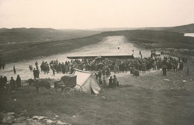 A black and white photograph of the Pride of Detroit surrounded by a large crowd on a clay airstrip, its wing visible over the crowd. A horse and buggy is parked next to a tent, at the foot of the airstrip.