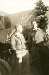 A black and white photograph of James Medcalf and Terrence Tully in aviator uniforms standing in front of the Sir John Carling aircraft, between the cockpit and propeller.