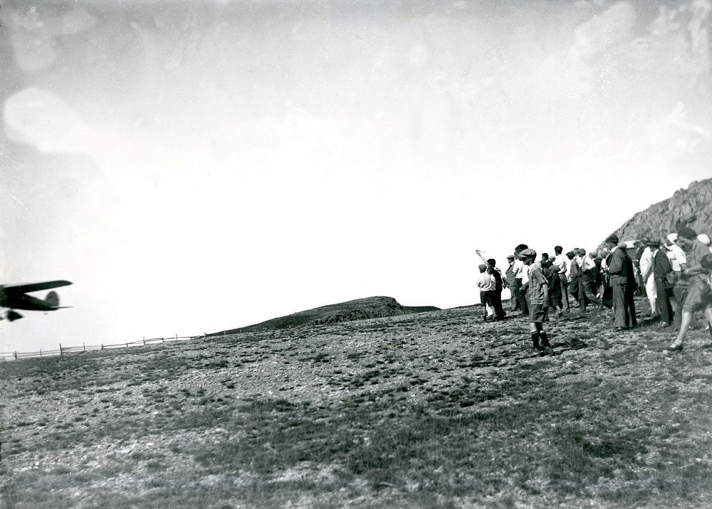 A black and white photo of Amelia Earhart's Lockheed Vega as it departs Harbour Grace airstrip. Half of the plane is visible as it takes off, approximately 30 feet in the air. A crowd is gathered to the right of the photo to watch her takeoff.