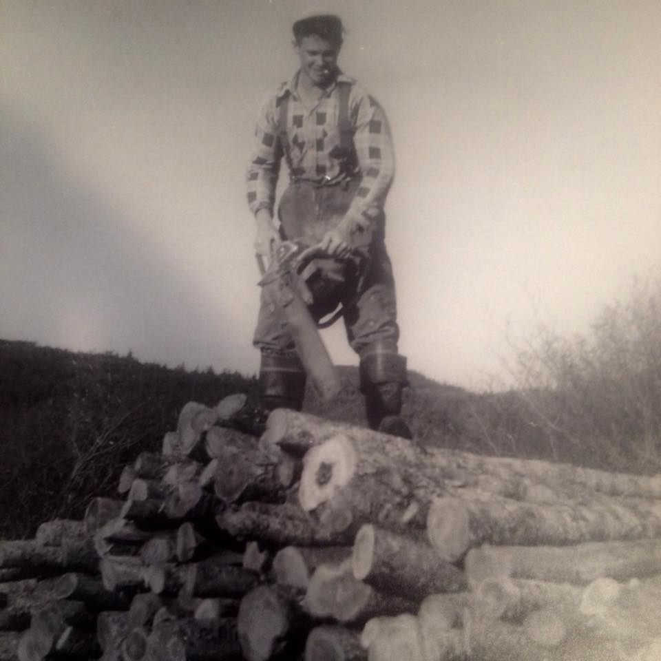 Wilson King stands on top of a pile of logs with a chain saw in hand.