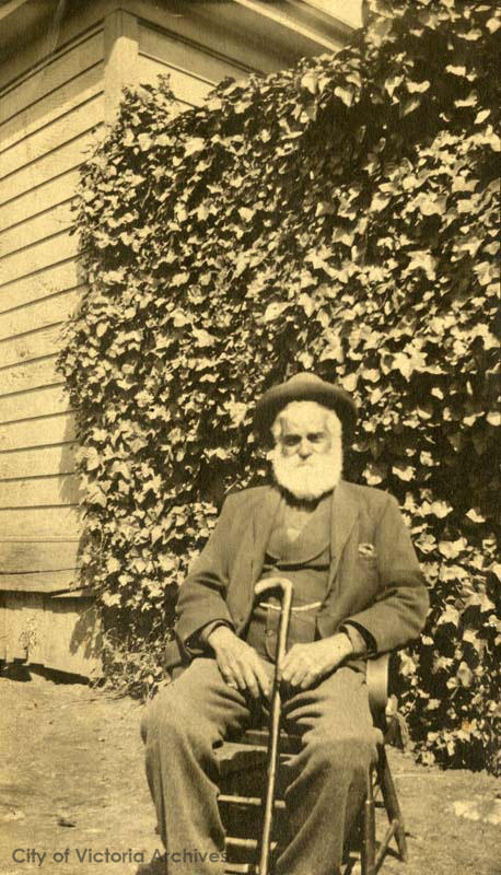 Sepia toned photo, man in senior years sitting in a chair with a cane leaning against his leg. In the background is a house and ivy hedge.