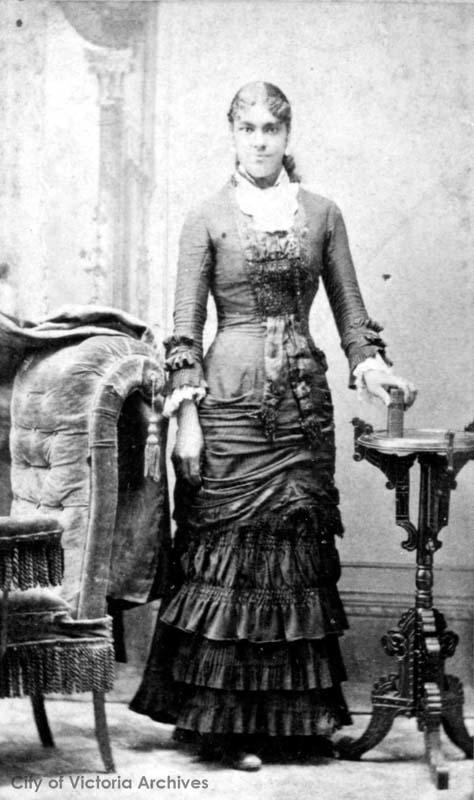 black and white photo, young adult female standing next to a chair posing for the camera with a slight smile; wearing a long formal dress, with a white under-blouse, ruffled at the neck and cuffs, long hair in a ponytail