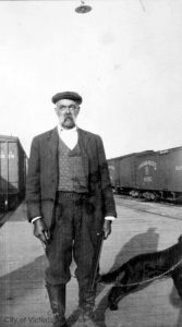 photo en noir et blanc d’un homme adulte portant un béret et un costume, debout sur le quai en bois d’une gare. On aperçoit des wagons de train derrière et à côté de lui. Il tient un fusil dans la main gauche, une laisse dans la main droite avec un chien près de lui