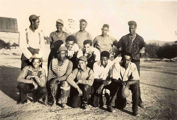 Sepia tone photo. 12 teenagers and young adults in three irregular rows, standing, crouching or kneeling. Dressed casually, happy, wearing a variety of clothing, hats and caps.