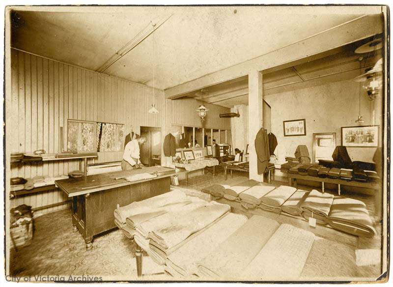 Black and white photo, inside a building there are several tables with many folded bolts of fabric on them. There is a counter with a man standing behind it.