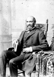 black and white professional studio portrait of middle-aged man, seated, dark hair and trimmed dark beard, wearing formal suit