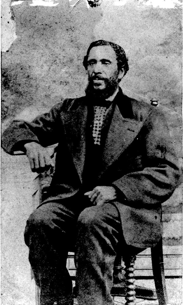 portrait studio professionnel en noir et blanc d’un homme d’âge moyen, assis, cheveux foncés et barbe taillée, portant un costume formel