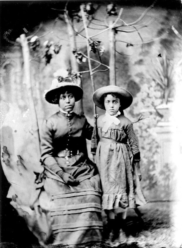 black and white professional portrait 2 sisters in colonial dresses wearing hats, girl age 23 left is seated, girl age 11 is standing, deciduous saplings in background