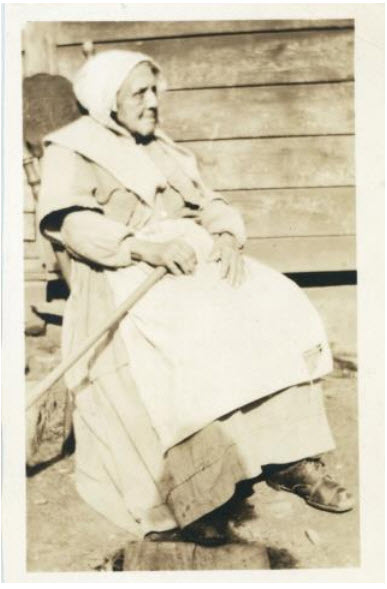 sepia photo, old woman wearing bonnet, long work dress, apron sitting in rocking chair on porch holding rake across lap in left hand.