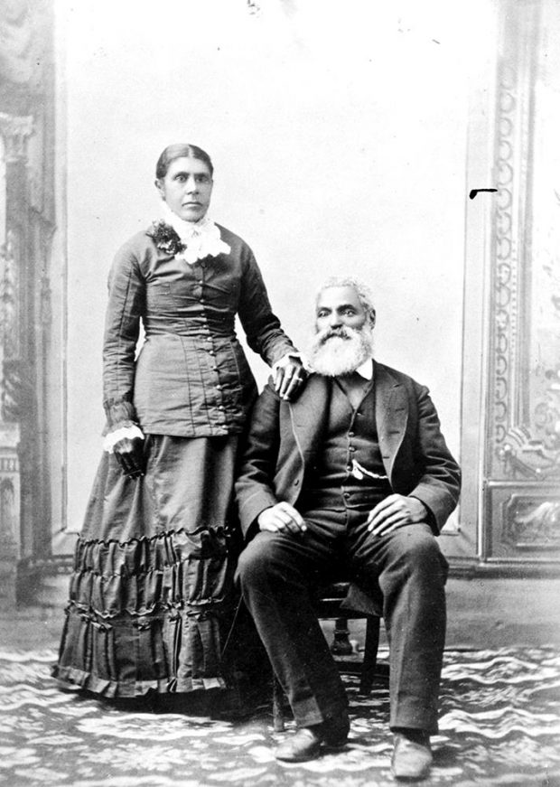 Black and white professional studio portrait woman standing, man seated. Nancy's hair parted in the centre, pulled back into a bun. Her right hand rests on Charles shoulder. Charles, seated to her right, with his right foot slightly forward wearing a dark suit and vest, his watch chain is visible from his vest pocket. Charles hair is white; he has a moustache and full beard.