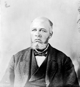 portrait en noir et blanc, homme âgé, chauve avec barbe courte et favoris sel et poivre, vêtu d’un costume avec veste, chemise blanche et nœud papillon