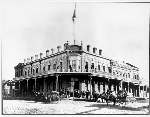 multi-windowed 2 story brick building, front door facing the street corner with 2nd floor balcony along both sides with railing. Roof shows a dozen chimneys. Patrons stand on lower verandah and several hacks are waiting on the street.