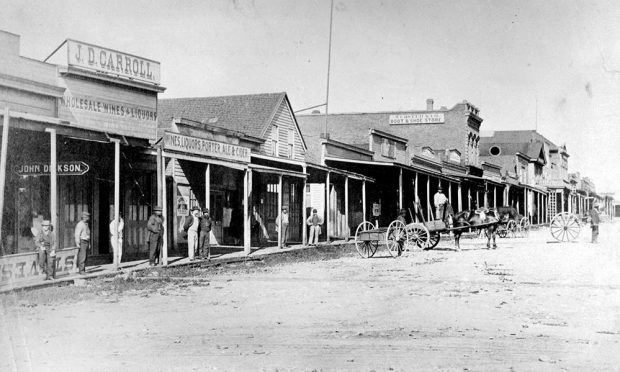 wide dirt street lined on both sides with businesses, constructed of wood and brick of various lengths and heights and roof types. People are walking or standing at storefronts, horses and carts travel the streets.