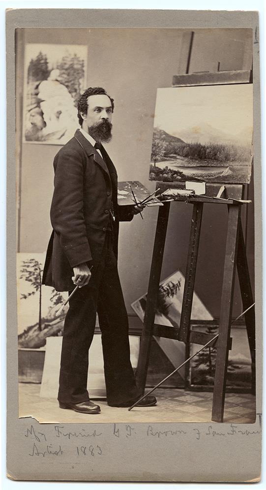 young bearded man standing at easel with paint brushes and palette in left hand and 1 brush in right hand