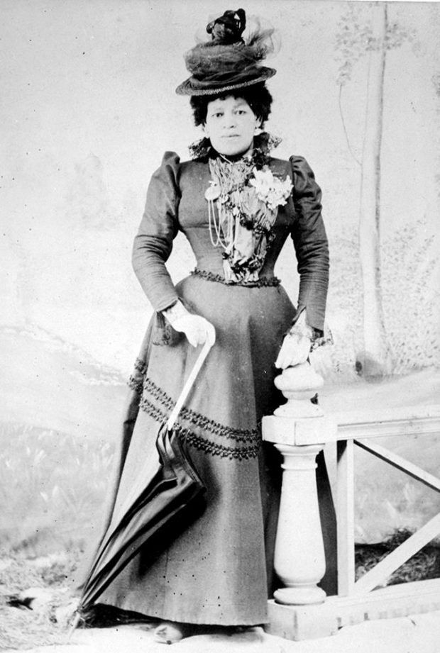 black and white professional studio photo of woman standing regally, leaning on closed umbrella; wearing tiered Victoria hat adorned with lace and flowers, long flared dark dress, ruffles and piping adorn the neck and bodice