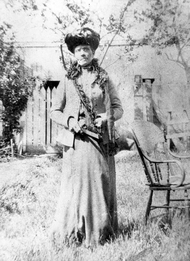 black and white professional photo of woman standing regally in a garden, wearing tiered Victoria hat adorned with lace and flowers, long flared light-colored dress, with hip-length suit jacket with ruffled lapel.