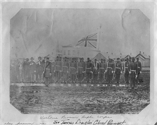 About 20 members of the Victoria Pioneer Rifles Corp stand in their ranks with the British flag in the background