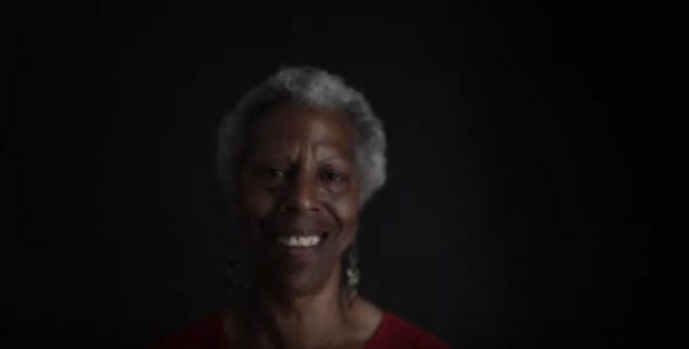 Portrait shot of slim, older woman with close-cropped afro hair style, smiling, in a studio, black background.