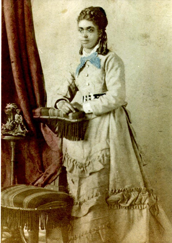 Hand tinted photo, young adult female standing next to a chair posing for the camera with a slight smile; wearing a long light-colored dress with wide belt, ruffles on the skirt. hair is in ringlets