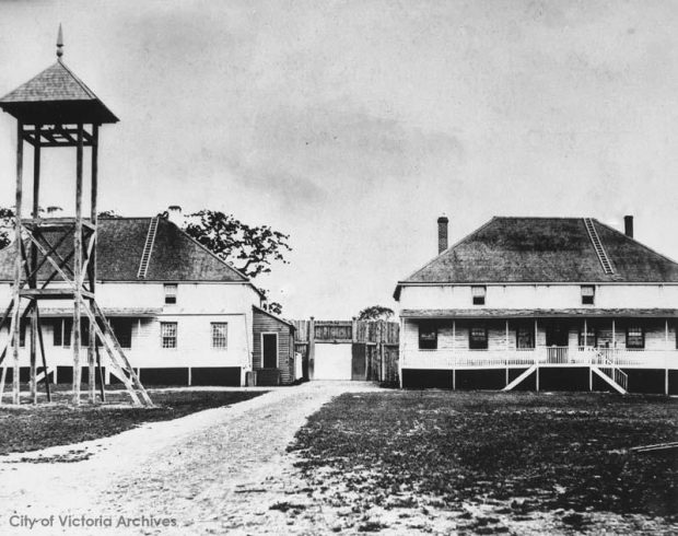 Black and white photo, a lookout tower stands before two large buildings. There is a gate between them with a pathway that goes between the buildings.