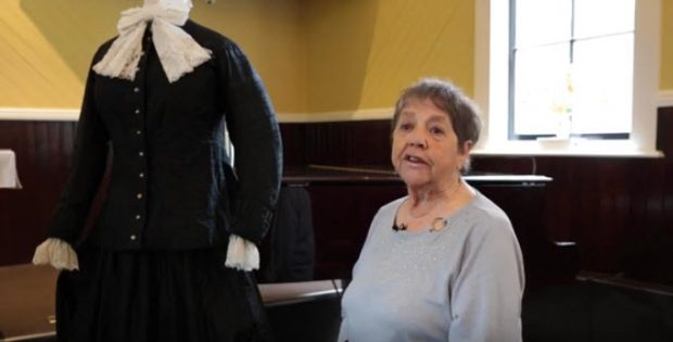 older woman sitting on piano bench in a church turned to the camera; mannequin with long black colonial dress to her right