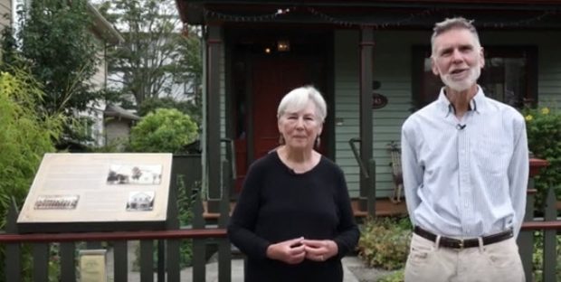 older couple standing on sidewalk in front of house. woman's hands are clasped in front, man is standing with hands behind back