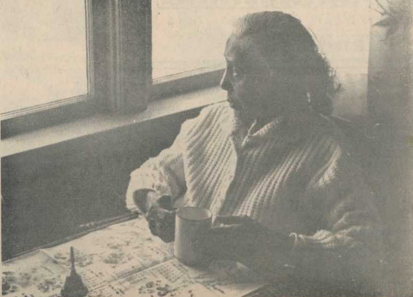 sepia photo, old woman seated at table with cup of tea and newspaper open on table, looking out a window