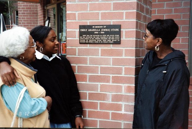 3 women of varying ages viewing a plaque mounted on a brick wall.