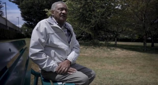 senior man with salt&pepper hair & moustache sitting on park bench in grassy park, turned to the camera. evergreen trees in background