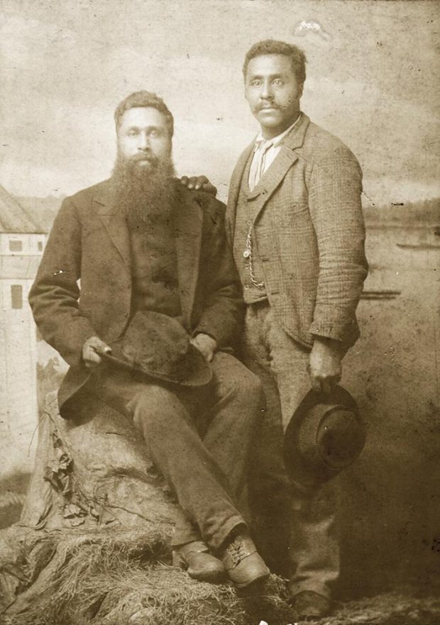 sepia toned photo, man seated on rock with full beard holding hat, man standing beside him, clean-shaven holding hat, seascape background