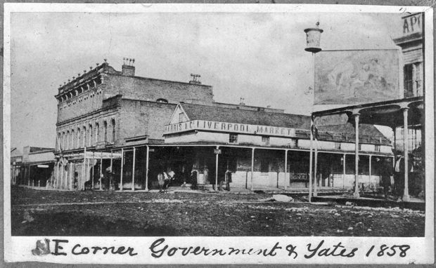 Text at bottom says NE corner Government & Yates 1858 intersection of 2 dirt streets with businesses buildings constructed of wood and brick of various lengths and heights, and roof types. A few people and horses can be seen