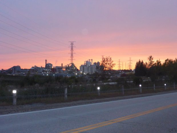 A colour photograph of a number of industrial buildings and set against a colourful sky and at a distance from a road