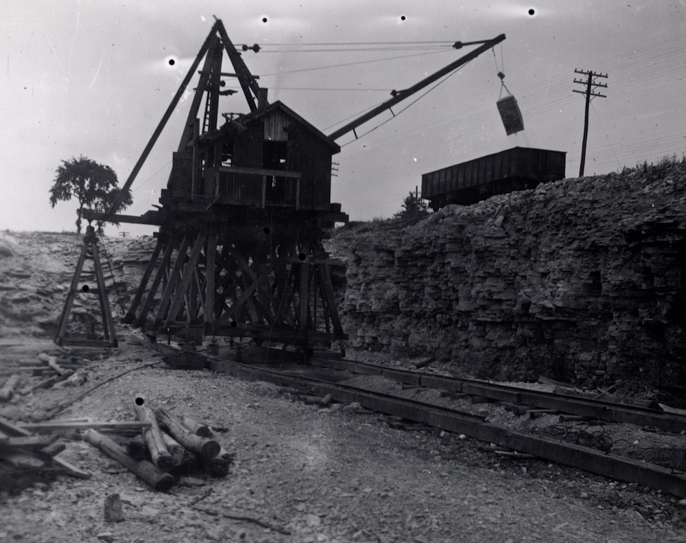 Black and white image of a piece of equipment in a quarry pit on tracks - The device is hoisting a load of supplies onto a railway cart at the edge of the quarry.