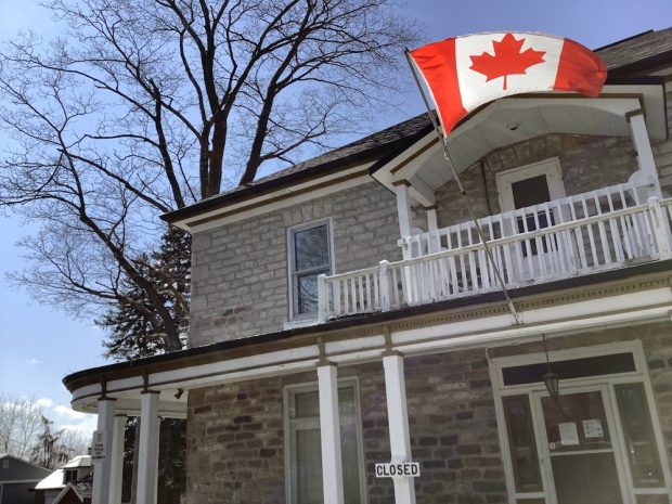 A partial facade of a stone building showing a white porch and a second-floor balcony - A Canadian flag flies atop its front door.
