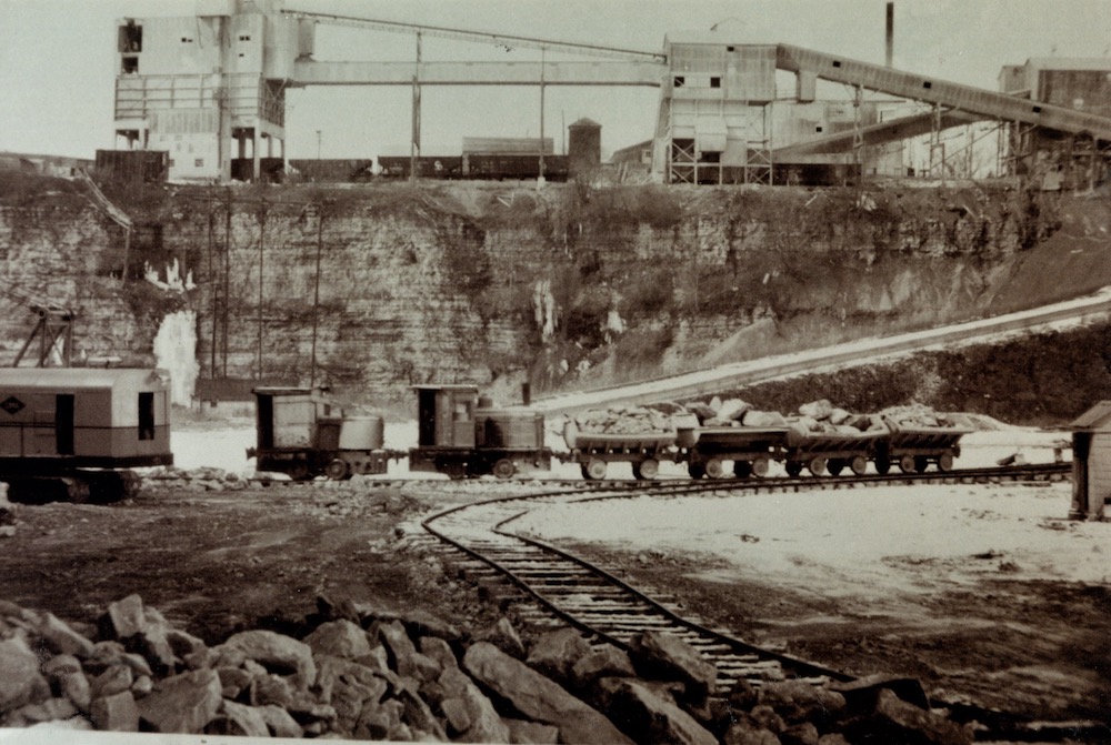 A small train and four carts are seen along a track inside a quarry pit. In the background are several industrial structures.