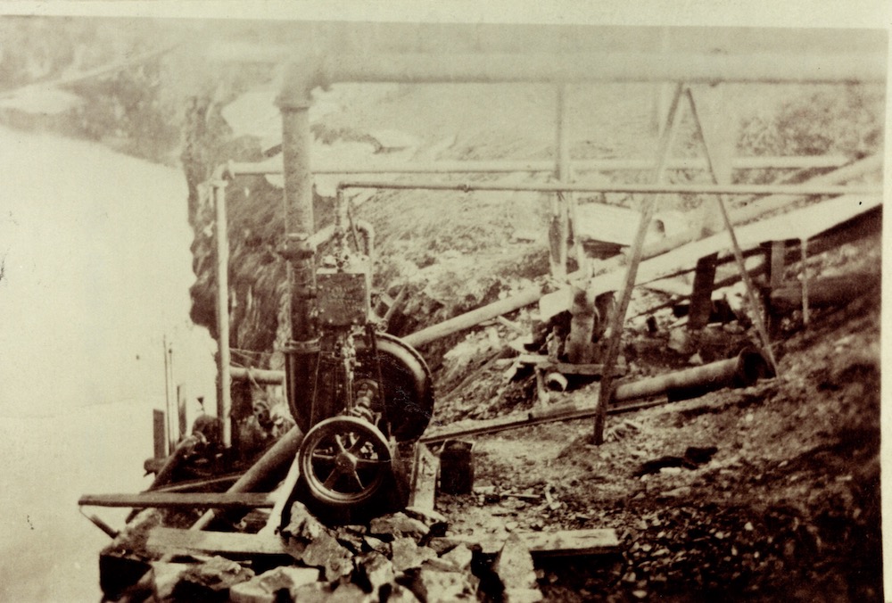A black-and-white photograph of a water pump with attached pipes on the edge of a quarry pit