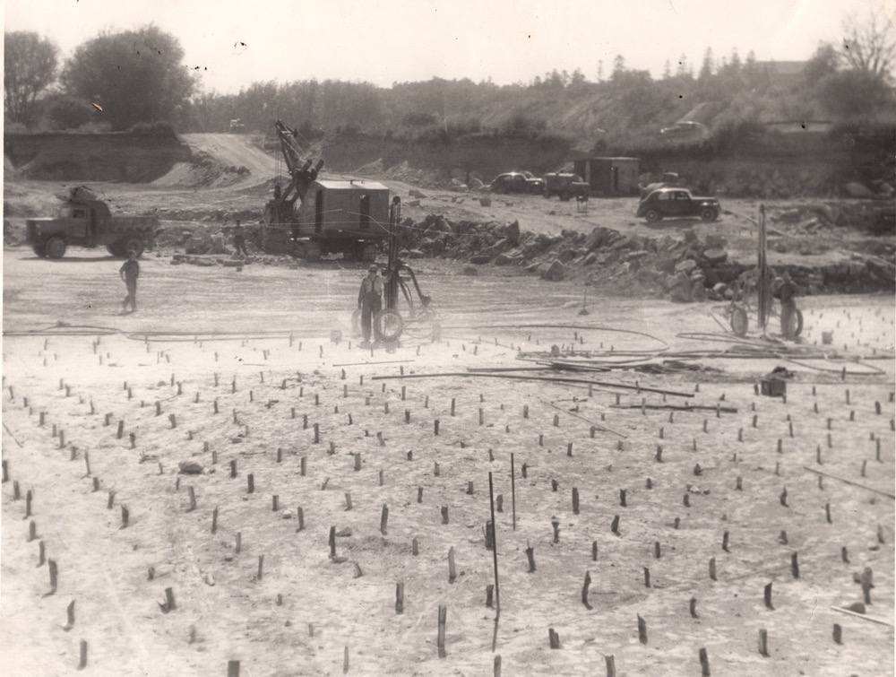 Sticks of dynamite pop out of the ground in rows. In the background are cars, heavy machinery and piles of stone.
