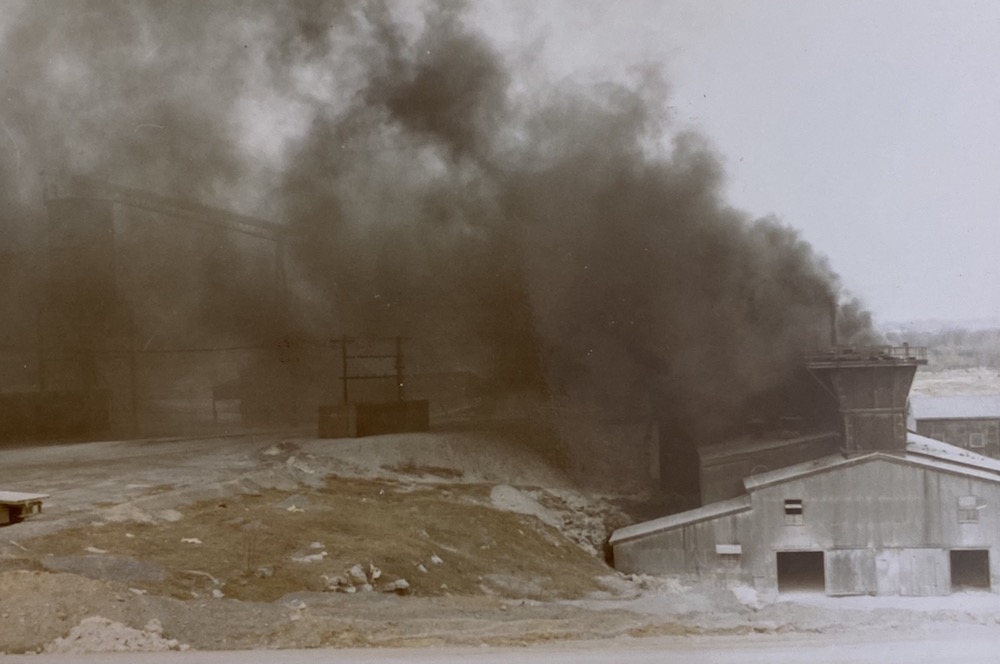 A cloud of dark smoke rises from an industrial building, obscuring the landscape and buildings in the distance.