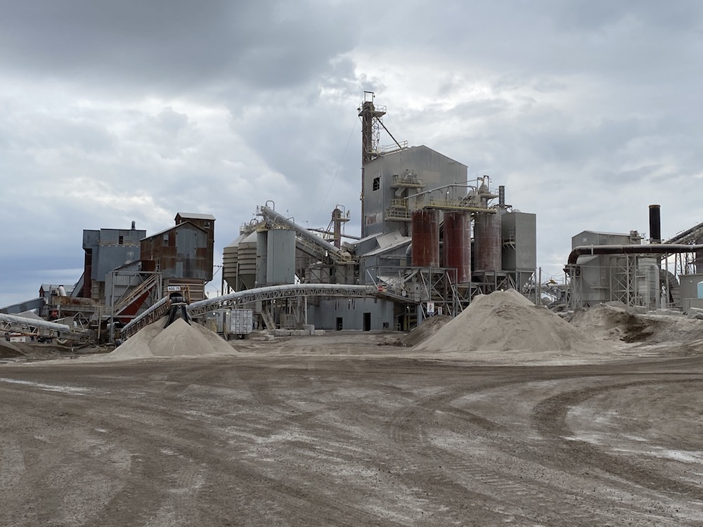 A colour image of several industrial buildings, silos, and gravel piles against a cloudy sky