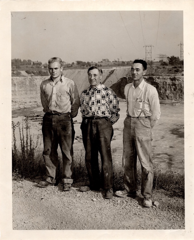Black and white photograph of three men standing together at the edge of a quarry pit.