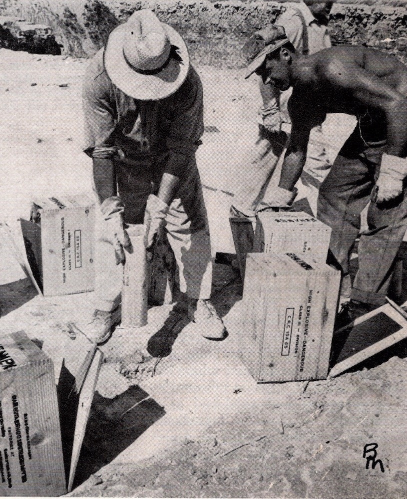 Two men hunch over boxes of dynamite and a drilled hole in the earth.