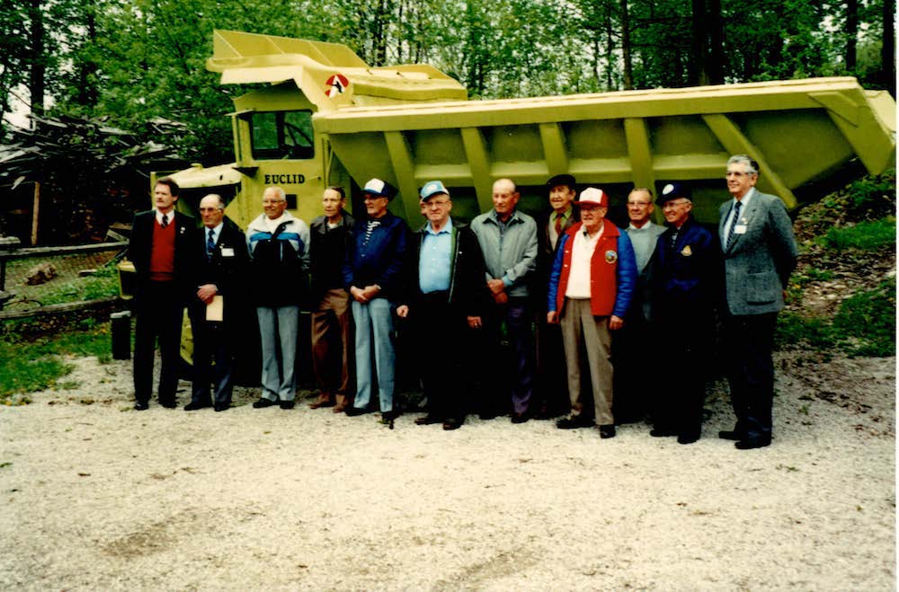 A group of 12 men stand in front of a green rock hauler truck