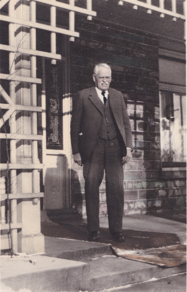 A man in a three-piece suit stands on a house's latticed porch squinting against the sun
