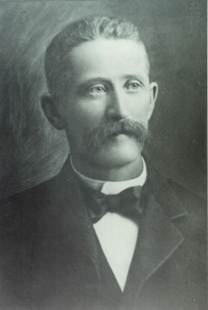 A black and white portrait of a man with a moustache wearing a suit