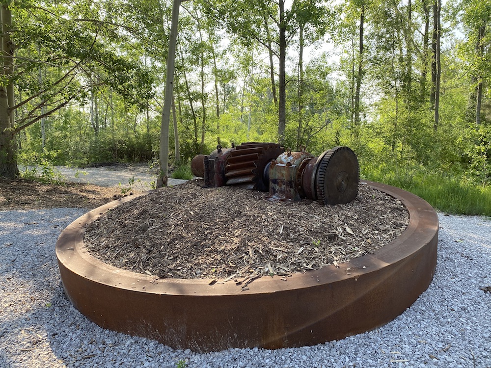 An image of a rusted gear set on a pile of mulch surrounded by a circular, metal border