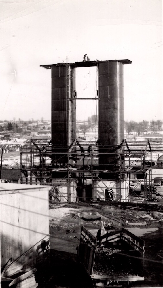 Two people work on a platform between two cylindrical structures that rise from a building frame