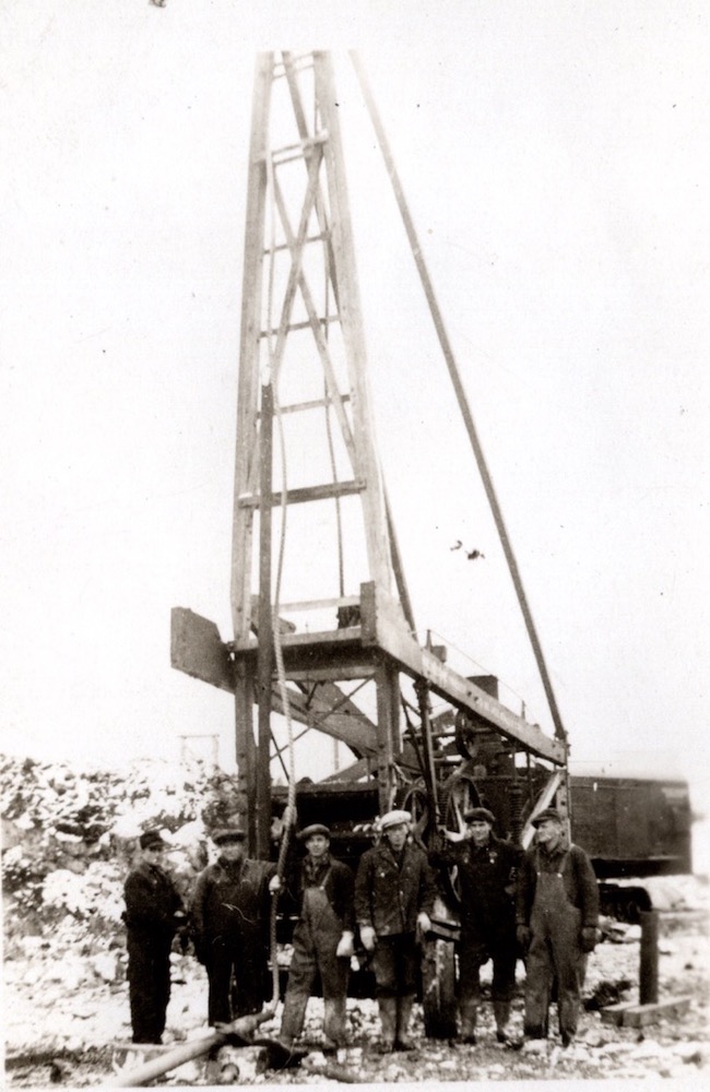 A black and white photograph of six men standing in front of a tall drill rig in jackets, suspenders and hats