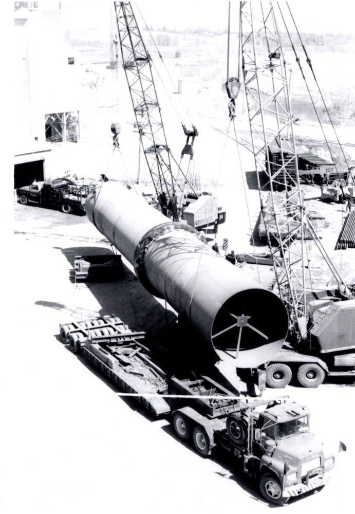 A large cylindrical part of a rotary kiln is hoisted off a flatbed truck by a set of cranes