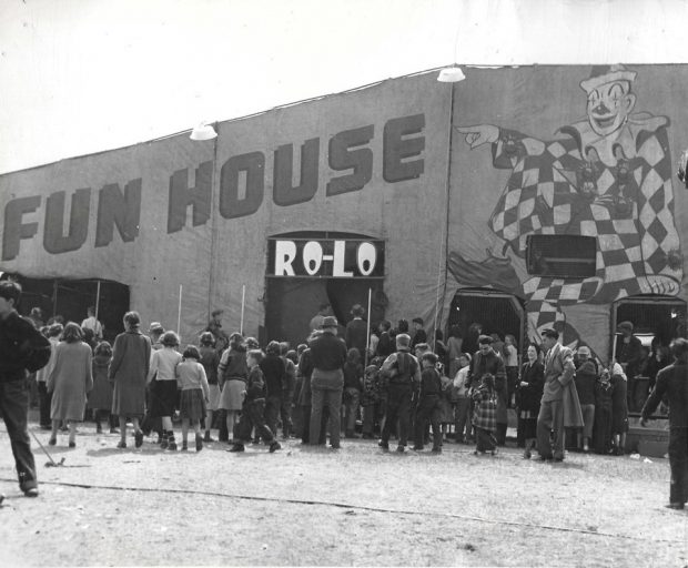 Black and white photo of people lined up to get into the Fun House, a large structure with canvas walls on which a large clown is drawn.