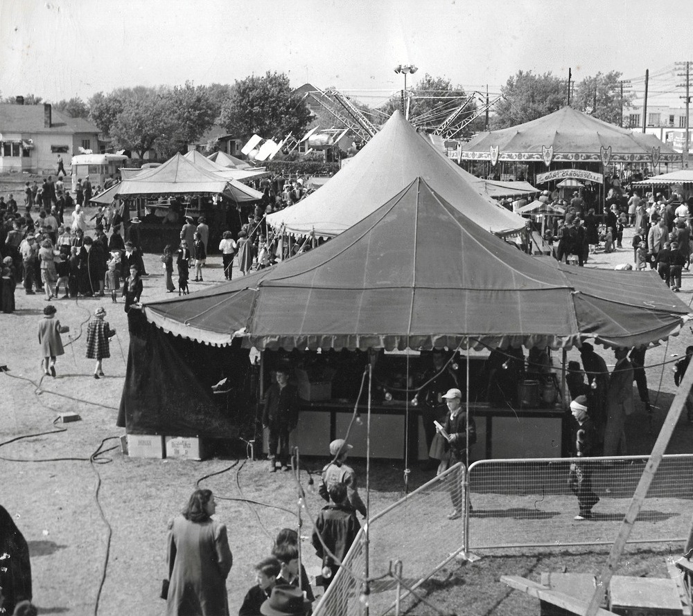 Black and white photo of crowds of people at a carnival midway with many tents set up and carousel in the background